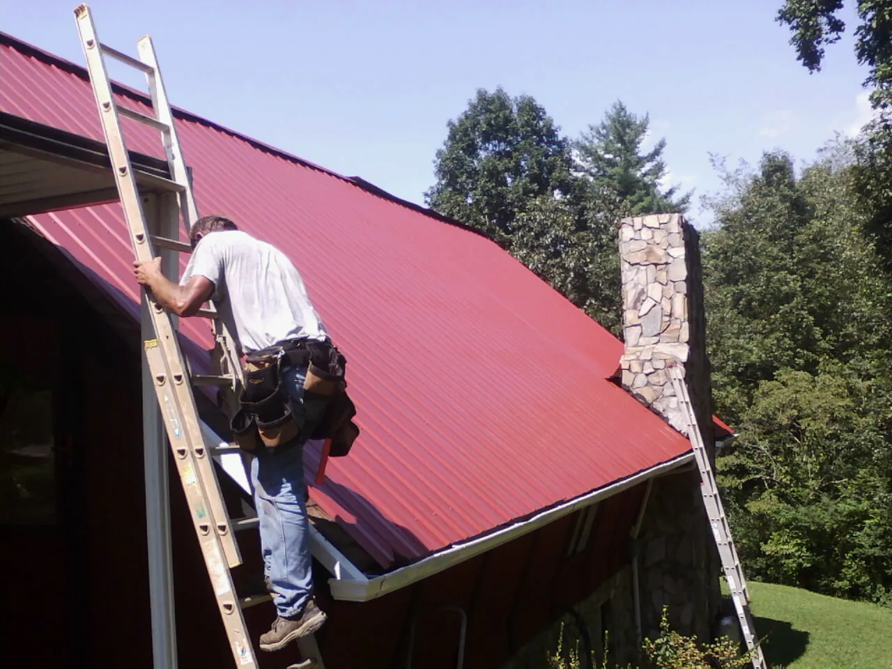Roofer on a ladder beside a red metal roof and stone chimney — Soffit & Fascia Repair by Roofing Brothers in Cougar